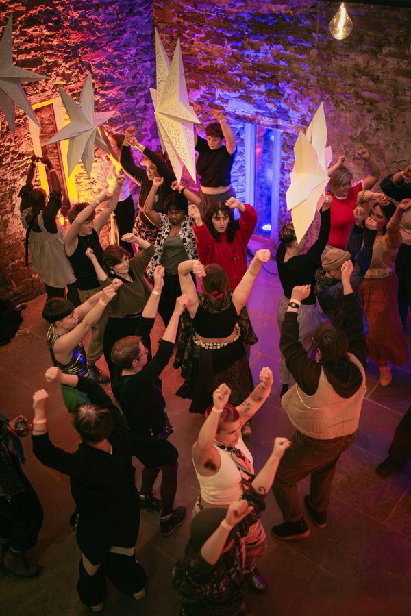 A group of about twenty people dances with their arms raised inside a stone-walled room lit in pink, purple, and amber tones. Large white star-shaped decorations hang overhead, adding to the festive atmosphere.