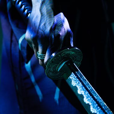Close-up of a hand gripping a decorated katana handle, the blade gleaming under blue light.