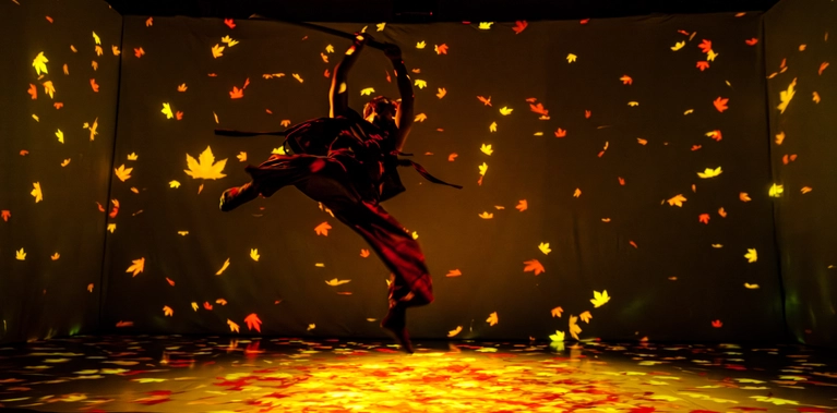 A man suspended mid-jump in a martial arts pose, holding a sword overhead, surrounded by falling leaves.