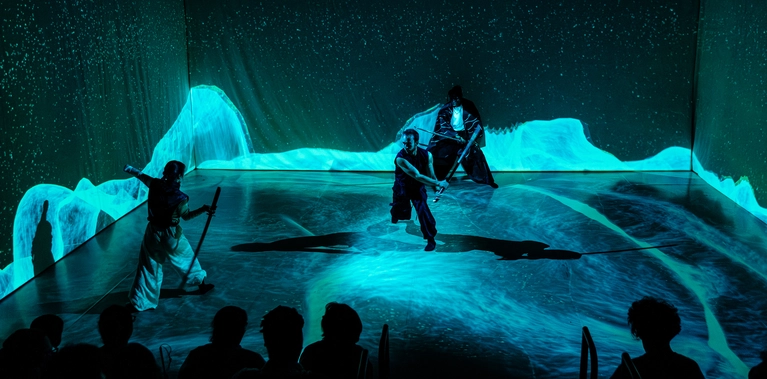 Three adults are engaged in a swordfight. A cold blue light casts abstract, contrasting patterns across the dark floor and background, which encloses them like four walls. Scattered points of light evoke a starry sky. Silhouetted figures in the foreground and visible fabric folds in the backdrop imply a theatre setting.