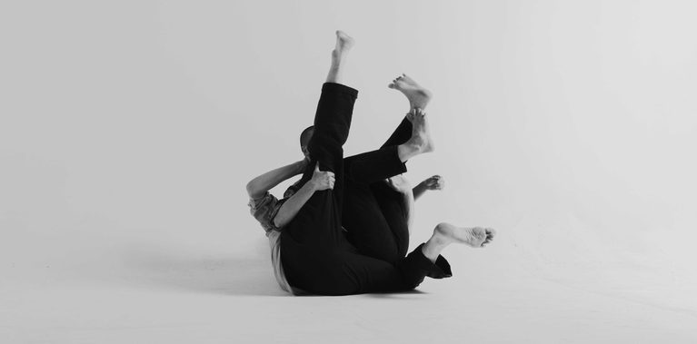 In a black-and-white studio image, two performers lie intertwined on the floor with overlapping legs and arms extending upward and outward against a plain background.