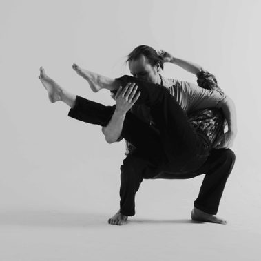 In a black-and-white studio portrait, one performer crouches while supporting another across their lap and shoulders in a complex partnered hold.