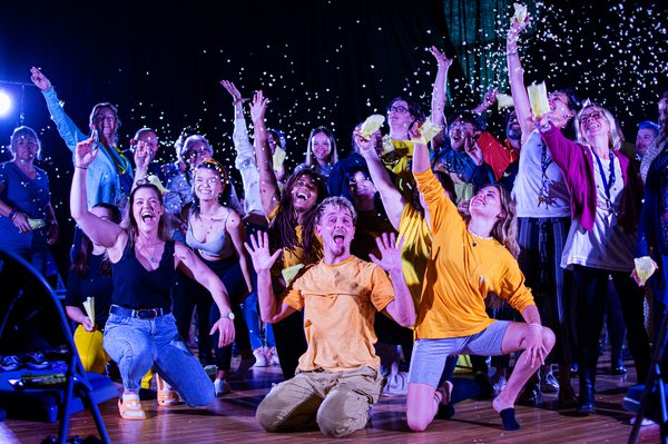 aKa Dance performers and the audience are all together in a large group, cheering and throwing confetti in the air. The performers are kneeling on the floor, whilst the audience are stood behind them.