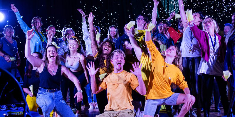 aKa Dance performers and the audience are all together in a large group, cheering and throwing confetti in the air. The performers are kneeling on the floor, whilst the audience are stood behind them.