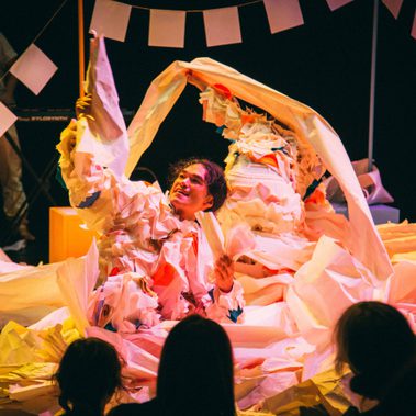 A performer is sat amidst a sea of paper, holding two pieces of paper in their hand and looking in excitement or amazement.