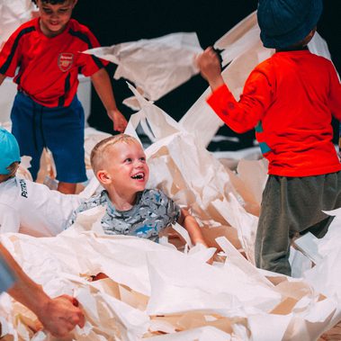 A group of children are joyfully playing in a sea of paper. Some are lying in the papers, others are seen holding or throwing sheets in to the air.