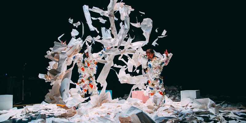 A photo of two performers dressed in scrunched up paper costumes, jumping and throwing paper up in the air.