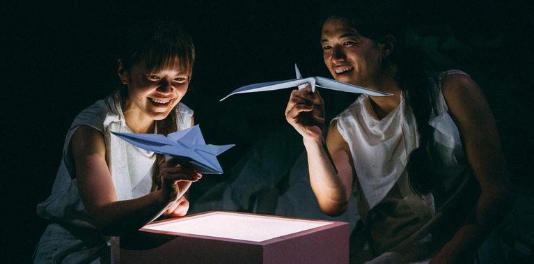 Two performers are sat around a square plinth which emits a soft light form the top. Both performers are holding paper airplanes and are smiling.