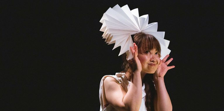 A performer holds a fanned paper circle around their head and smiles.
