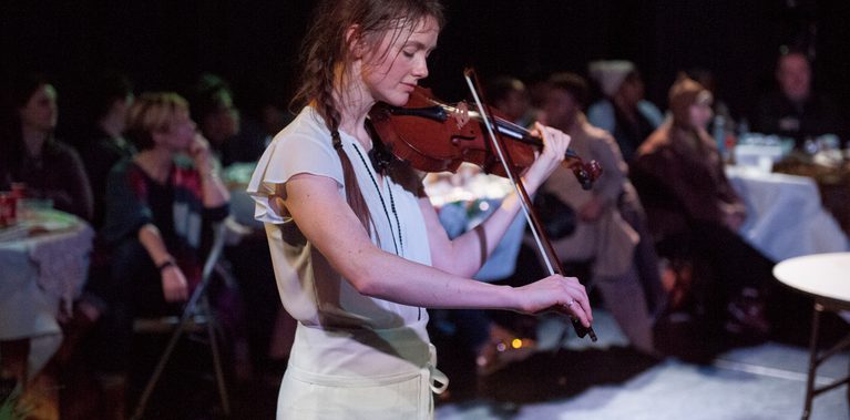 A female performer is captured playing the violin, their head bowed and eyes closed, as if lost in a moment of calm and focus. Begin them, and audience seated at dinner tables watches on.