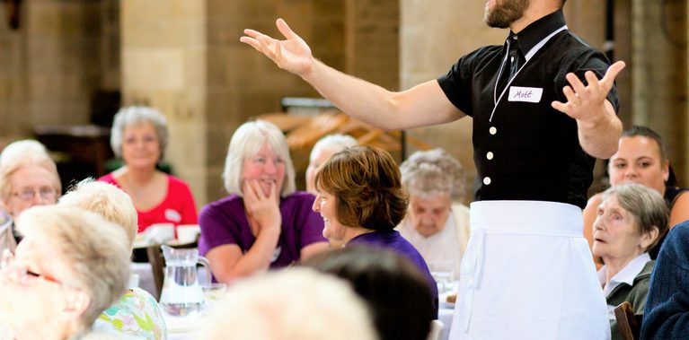 A performer, dressed as a waiter, stands with arms spread out, in the midst of either singing or talking with the audience. He is surrounded by an audience seated at tables, who look on with amusement.