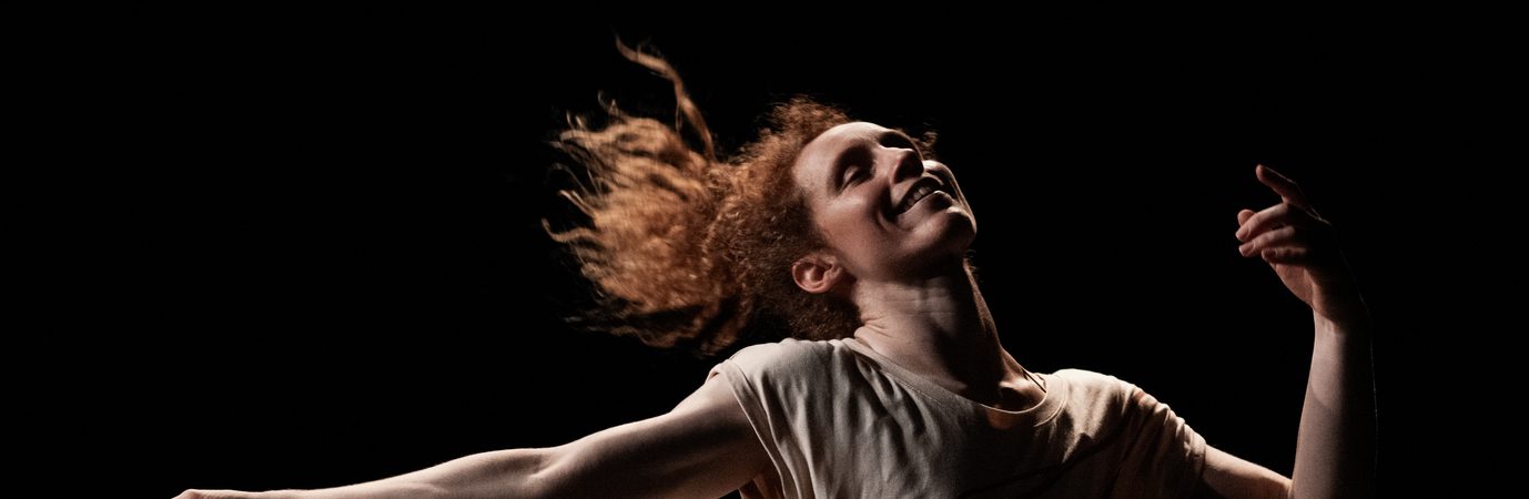 The performer, Nikki Rummer, is photographed mid-dance. Her arms are spread out and her legs are planted wide apart. She is smiling and appears to be liberated through the dance.