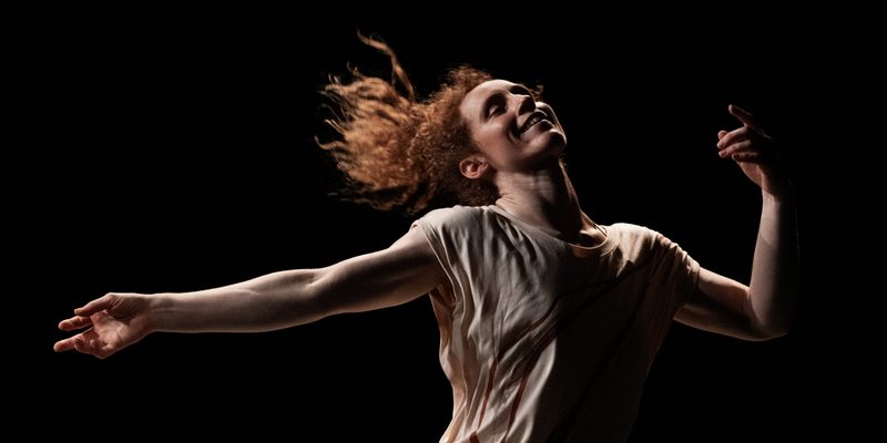 The performer, Nikki Rummer, is photographed mid-dance. Her arms are spread out and her legs are planted wide apart. She is smiling and appears to be liberated through the dance.