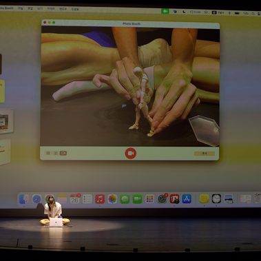 A performer sits on stage with a laptop, as a projected webcam feed shows hands manipulating a small figurine, blurring the lines between digital and physical movement.