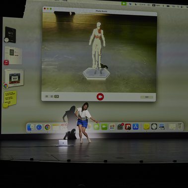 A performer moves in front of a giant projection screen, blurring the boundary between reality and the digital world. The scene prompts a reconsideration of the body through duality and reflection.