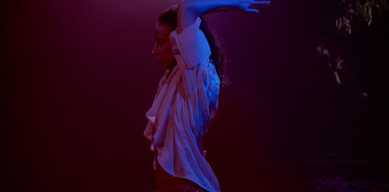 A profile of a person standing in a dimly lit space filled with red and purple light. They are wearing a white ruffled blouse and red pants, with their arms bent upward and hands crossed behind their head in a moment of reflection or preparation. A cool blue light hits their arms and hair, creating a sharp contrast against the warm, hazy background.