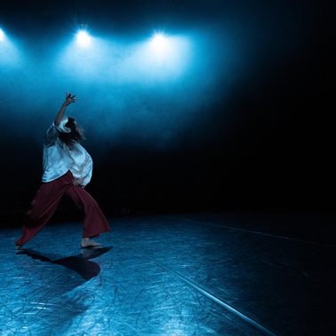 A dancer in a white top and red wide-leg pants performs on a dark stage. Three bright blue spotlights cut through a hazy atmosphere, illuminating the dancer and casting long shadows across the textured floor.