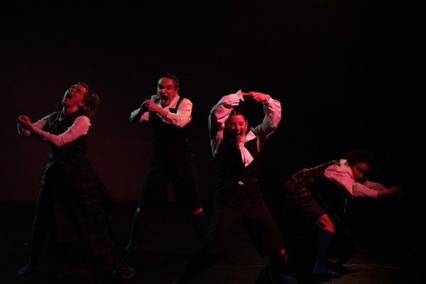 Four women in black dresses and white shirts dance expressively against a dark background