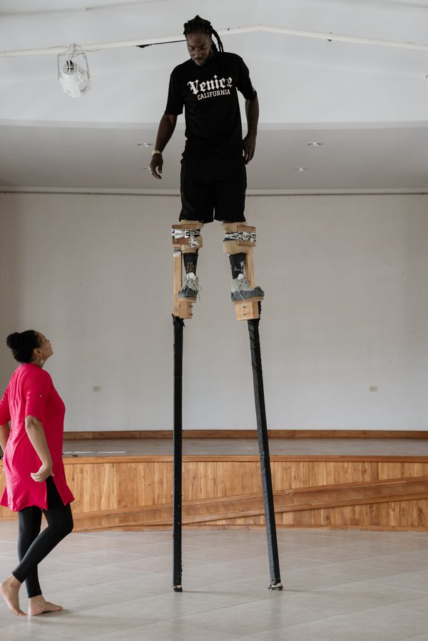 A person balances on very tall wooden stilts in a bright indoor hall while another person in a red tunic stands nearby, looking up at them. The stilt walker wears a black T-shirt and shorts, and the long stilts extend several feet above the floor, emphasizing their height.