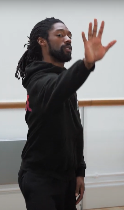 Person wearing a black hooded jumper and black trousers in a dance studio holding their hand out in front of them.