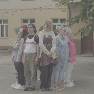 A group of people stands back-to-back in a circle facing outward in an outdoor courtyard, wearing pride-themed clothing and accessories.