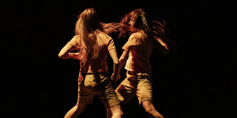 Two performers move in mirrored unison, their long dark hair flying around them. They are facing each other on a dark, glossy stage.