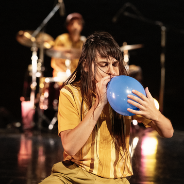 A dancer with long dark hair kneels and blows into a blue balloon, with a drummer visible in the blurred background.