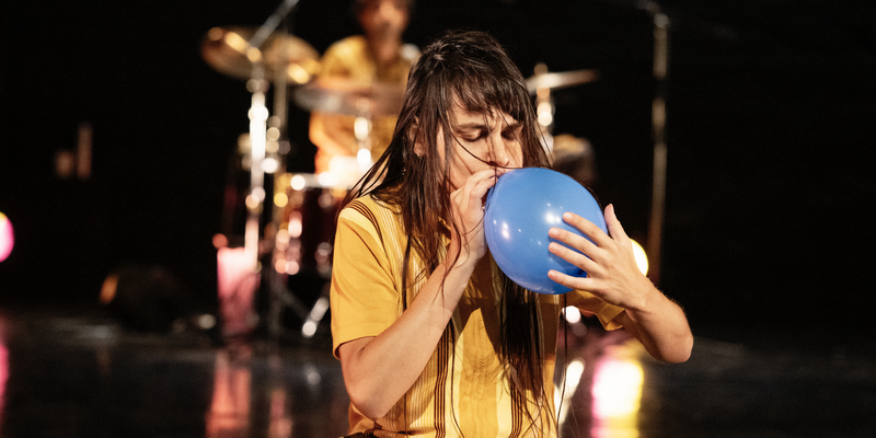 A dancer with long dark hair kneels and blows into a blue balloon, with a drummer visible in the blurred background.