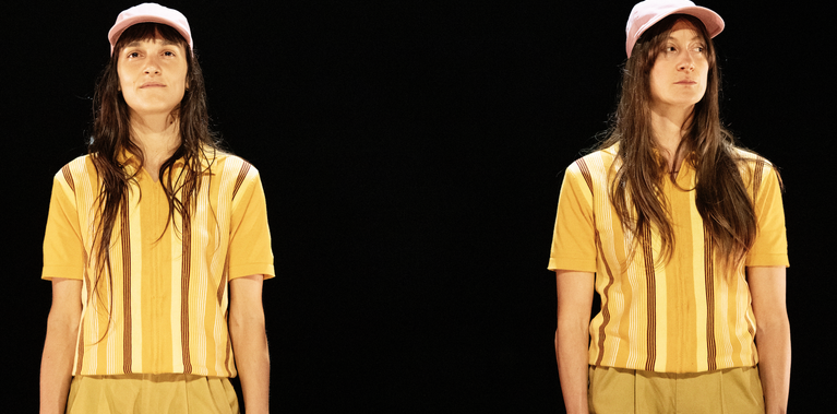 Two almost identical dancers with long dark hair wearing matching yellow striped shirts, tan shorts, and pink hats stand side-by-side, the performer on the left has a soft almost smile while the one on a right a more mischievous look. They stand together against against a black background.