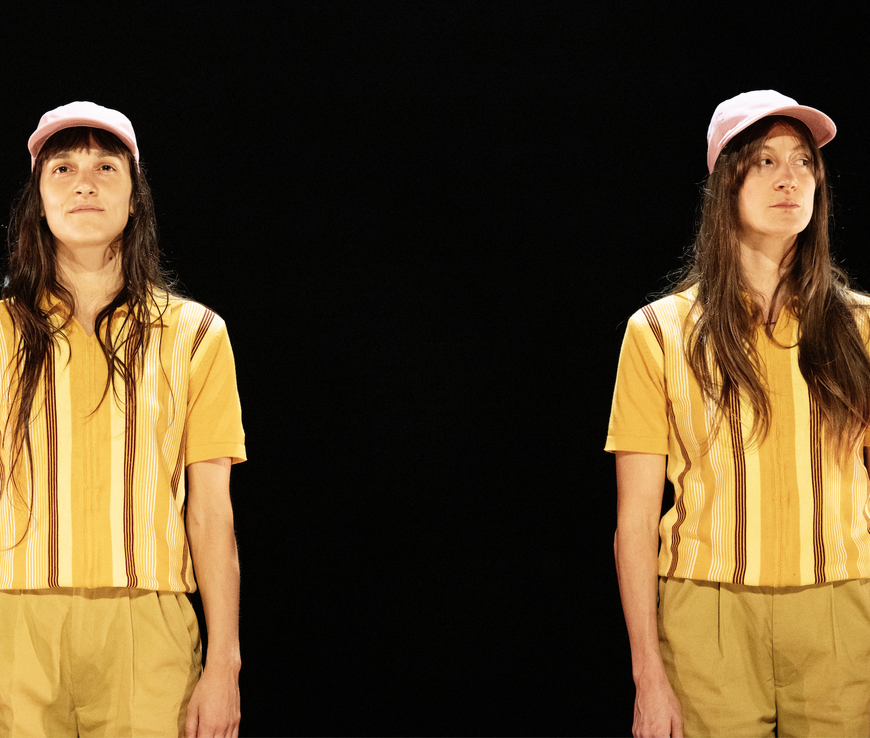 Two almost identical dancers with long dark hair wearing matching yellow striped shirts, tan shorts, and pink hats stand side-by-side, the performer on the left has a soft almost smile while the one on a right a more mischievous look. They stand together against against a black background.