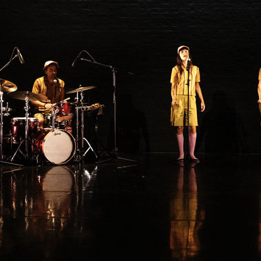 A drummer sits at a red drum kit on the left side of a dark, reflective stage. On the right, two performers stand with microphones, all three in matching outfits, yellow tops and bottoms as well as pink knee-high socks.