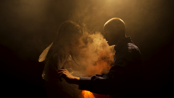 Two performers are captured in a dramatic, hazy scene lit by a warm, golden glow from above. The person on the right, who is bald and wearing a blue button-down shirt, stands in profile and appears to be speaking or reaching toward the woman. On the left, the other person's form is partially obscured by shadows and thick smoke or mist, creating a sense of mystery and tension between the two figures.