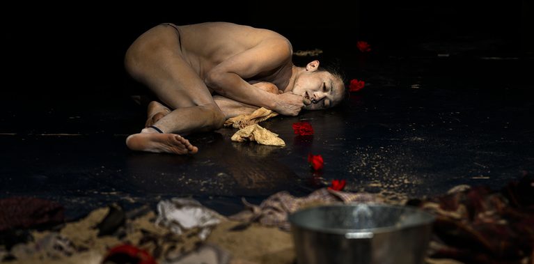A nude performer lies curled on the floor of a dark performance space amid scattered fabric and red flower petals, with a metal bucket in the foreground.
