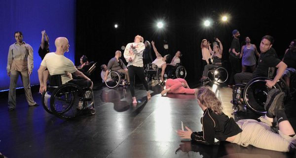 A group of performers spread across a theatre stage engages in a dance piece, with some standing, some seated in wheelchairs, and others moving or resting on the floor. Bright stage lights illuminate the scene as participants strike varied poses, creating a dynamic and inclusive performance.