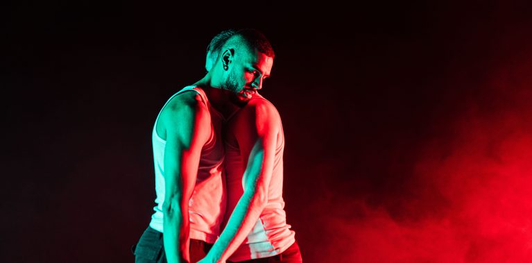 Two men lean into each other, wearing white vests, bathed in green and red light