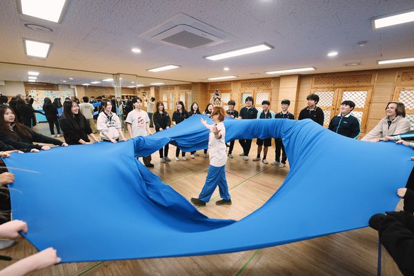 A group participates in physical activity indoors, holding a large blue fabric sheet in a circle. One person walks through the centre while others coordinate the movement of the fabric around them.