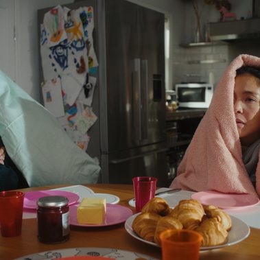 Two people sit side by side at a breakfast table in a kitchen, both wrapped in duvets. One person's face is completely obscured by the duvet, while the other’s face is partially visible, showing a tired, distant expression. The scene has a subdued, muted atmosphere, with a sense of quiet discomfort.