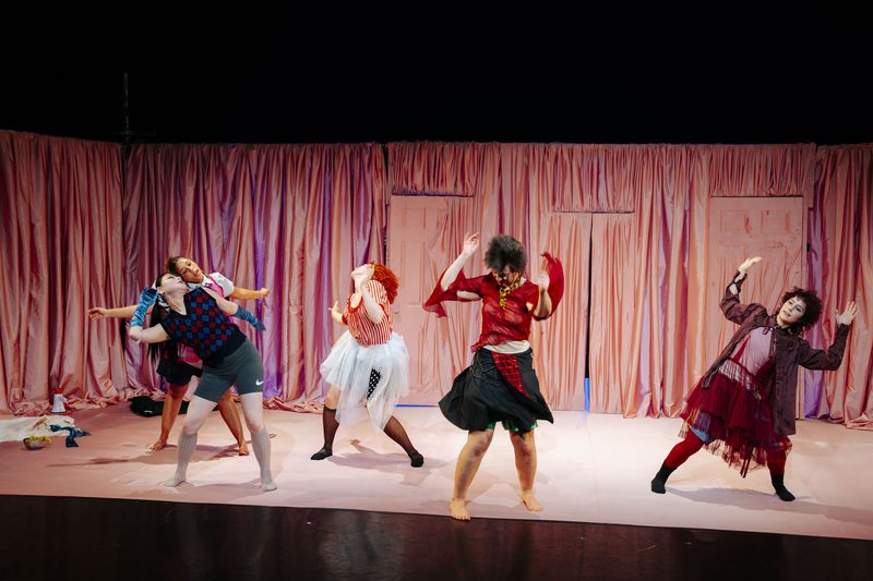 Five performers dance on a pink stage set with draped curtains and doorways, wearing colorful layered costumes and striking exaggerated poses under theatrical lighting.