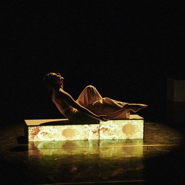 A performer reclines on a glowing rectangular platform in a dark theater, lit by warm stage light with one knee raised and their head tilted back.