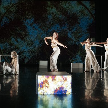 Five dancers in light costumes perform on a reflective dark stage with white blocks and a blue textured projection behind them, some standing with outstretched arms while others pose low near a metal frame.