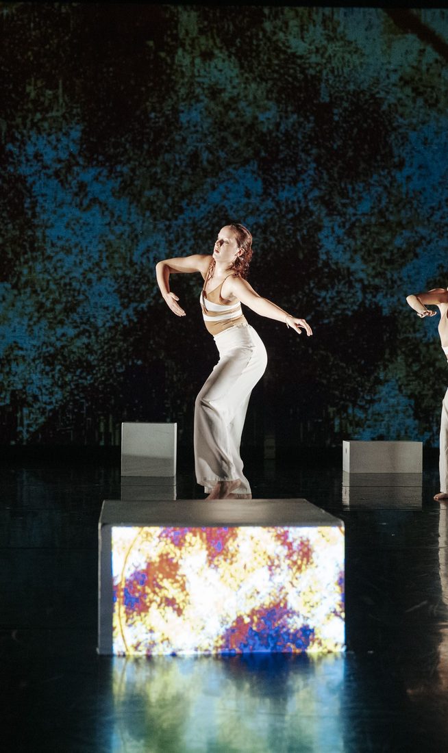 Five dancers in light costumes perform on a reflective dark stage with white blocks and a blue textured projection behind them, some standing with outstretched arms while others pose low near a metal frame.