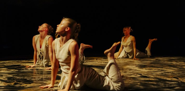 Three dancers in light-coloured costumes lie on their stomachs on a dark stage, arching their backs with legs bent as warm light reflects off the black floor, partially covered in sand.