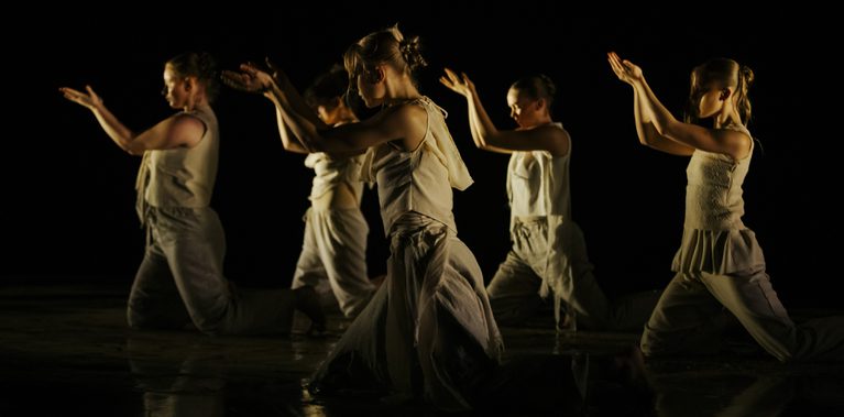 Five dancers in light-toned costumes kneel in a row on a dark stage, raising cupped hands in synchronized movement under warm, low lighting.