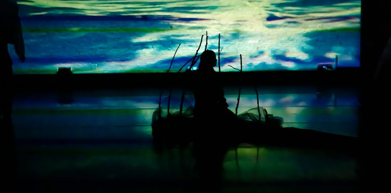A lone dancer sits in silhouette against a backdrop that looks like shimmering water. Surrounded by delicate stick-like props, the scene creates a serene, dreamlike atmosphere.