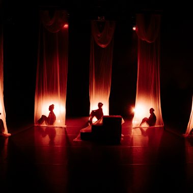 Five dancers are silhouetted behind tall, sheer curtains, each lit by a soft, warm glow.