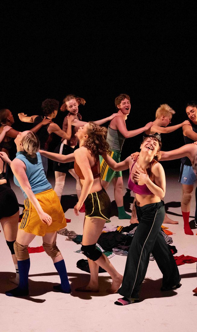 A group of dancers on stage in a circle dressed in colourful clothing and looking joyful.
