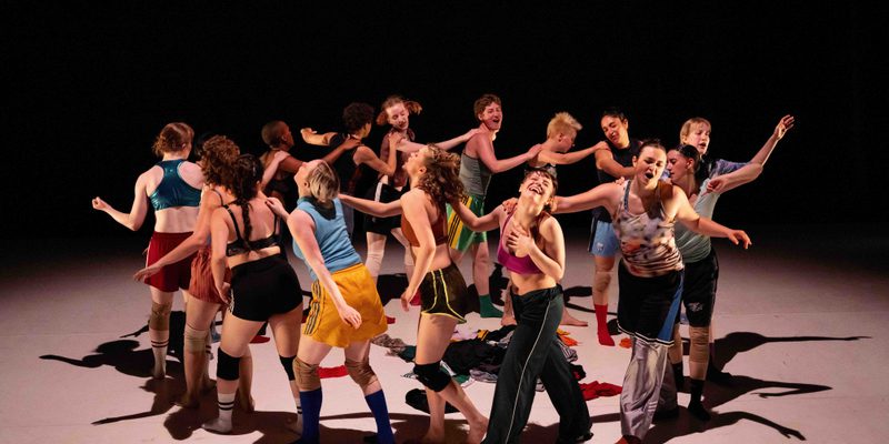 A group of dancers on stage in a circle dressed in colourful clothing and looking joyful.