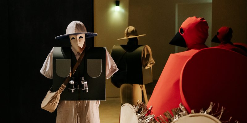 A live-installation featuring a masked bird-watcher with a pair of binoculars, and a performer wearing a large red bird costume