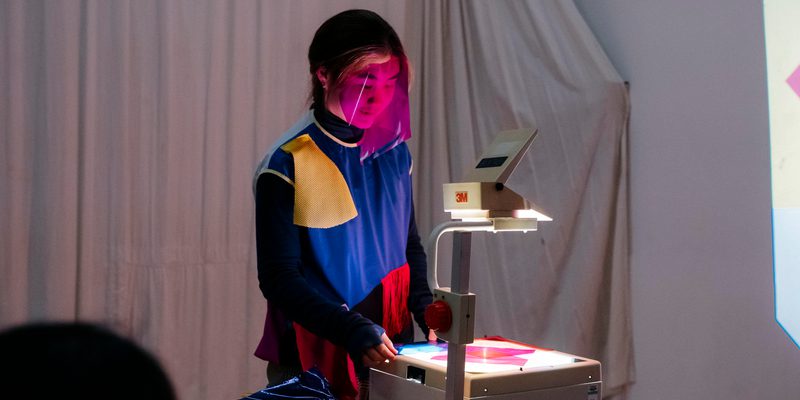 A performer stands over an overheard manual projector with a pink transparent gel masked over their face, projecting pink onto a white wall. They are wearing a black blue red and yellow colour blocked top, the wall behind them is covered with off white draped fabric.