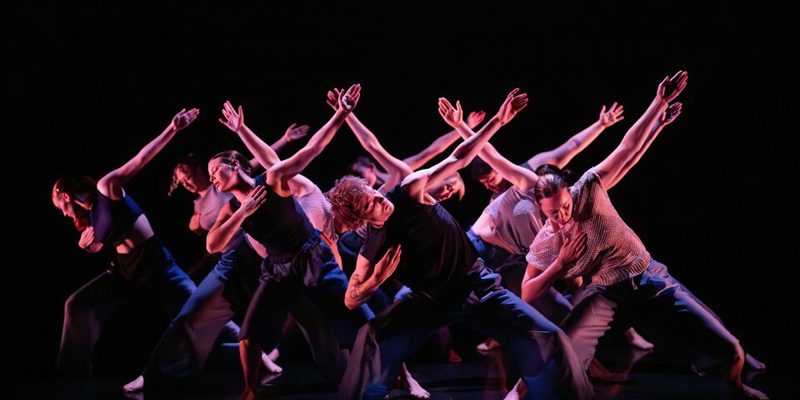 A group of dancers move in sync of stage underneath pink lighting. Their knees are bent, one arm is bent into their chest and the other arm is reaching out.